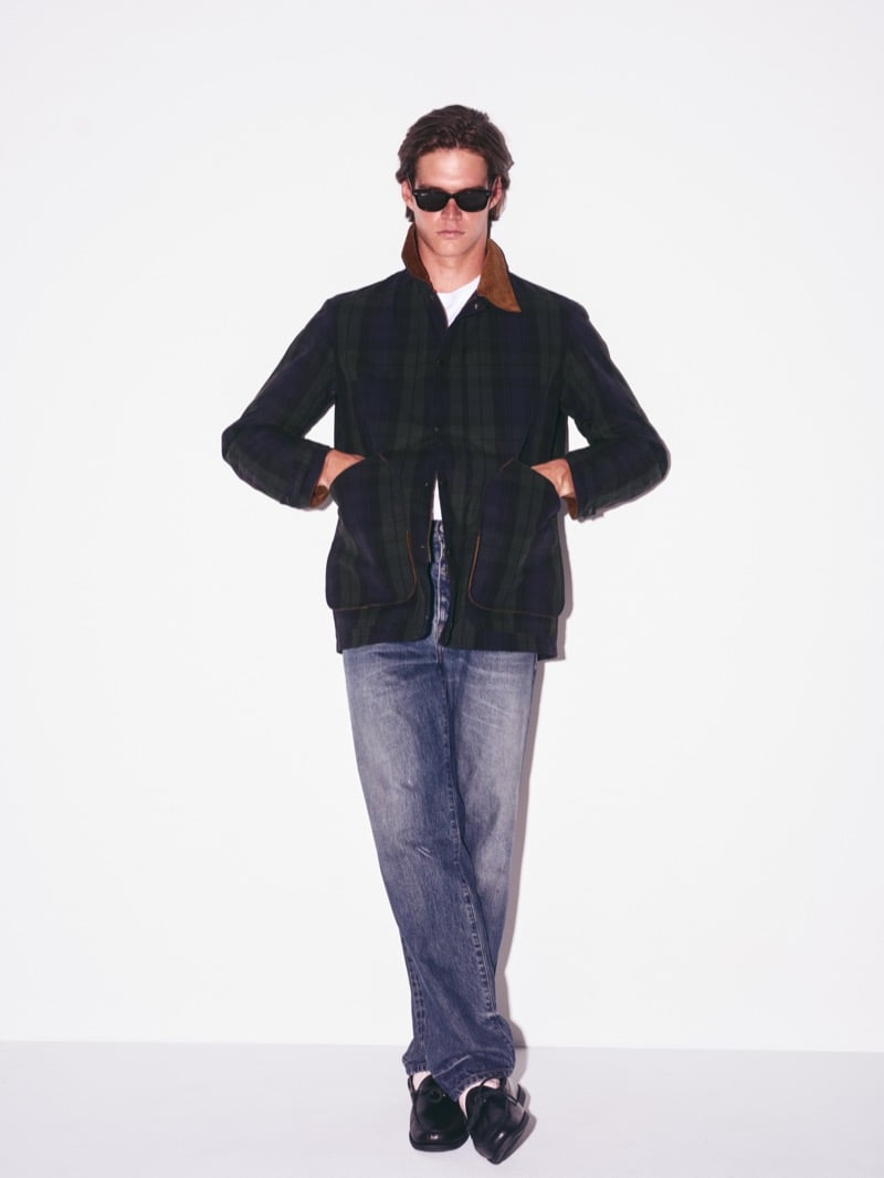 Drew Carter standing full-length in sunglasses, a Black Watch plaid barn jacket with large patch pockets, faded jeans, and loafers, hands tucked into the pockets