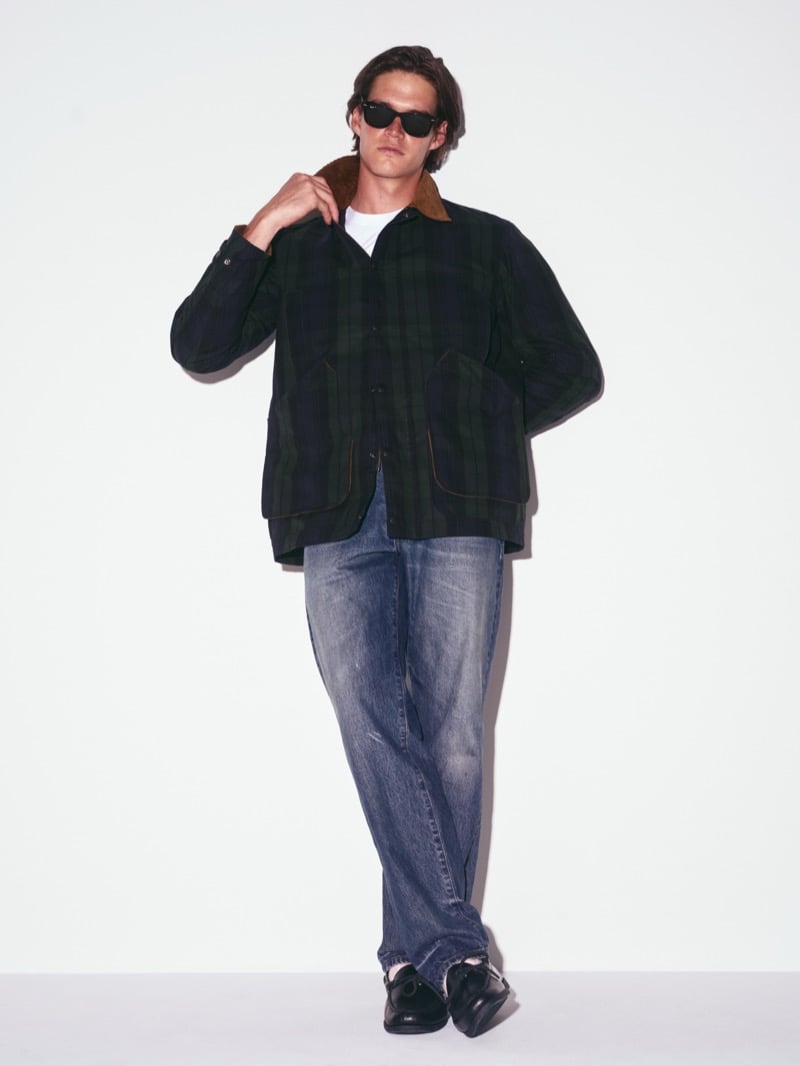 Drew Carter in sunglasses, a Black Watch plaid waxed barn jacket with a corduroy collar, faded straight-leg jeans, and loafers, one hand at his collar