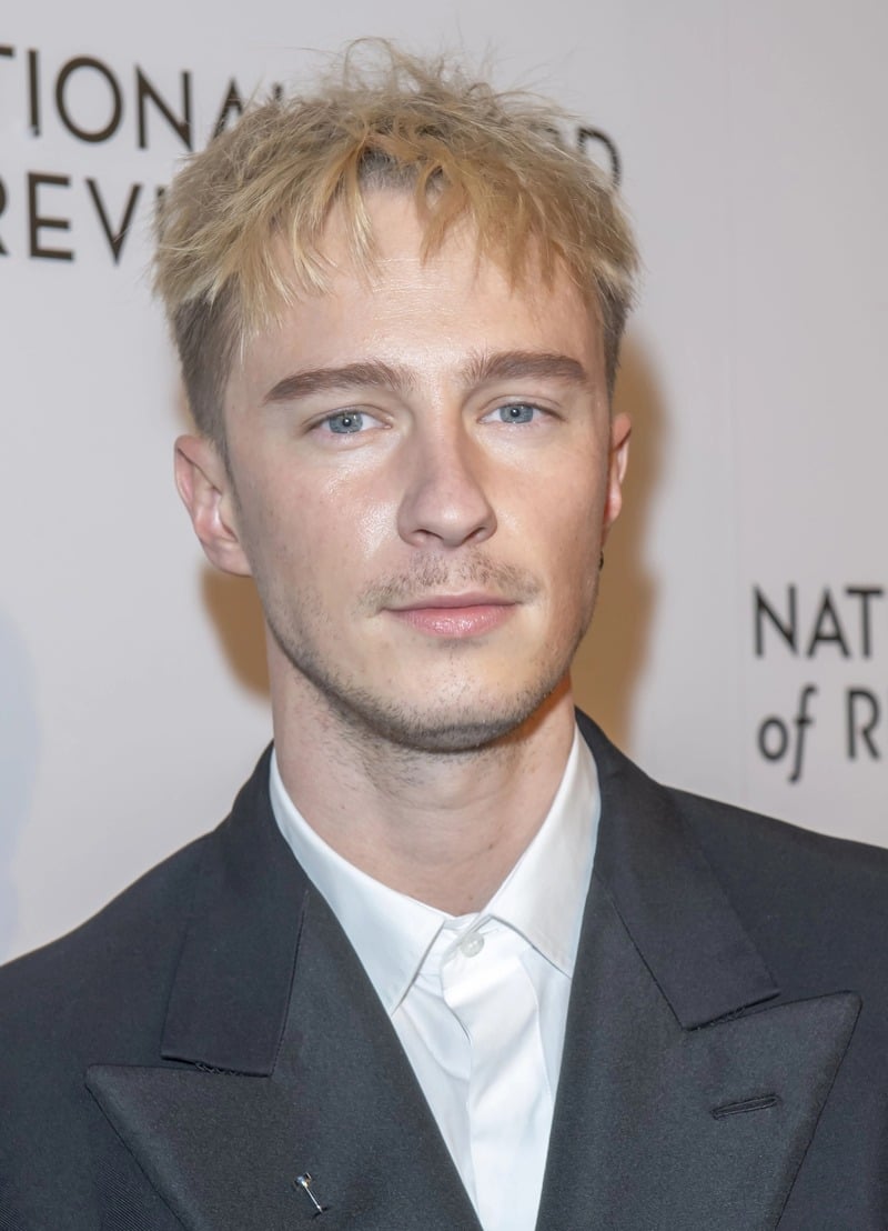 Drew Starkey with short, tousled blonde hair and light stubble, wearing a dark suit over a white buttoned shirt.