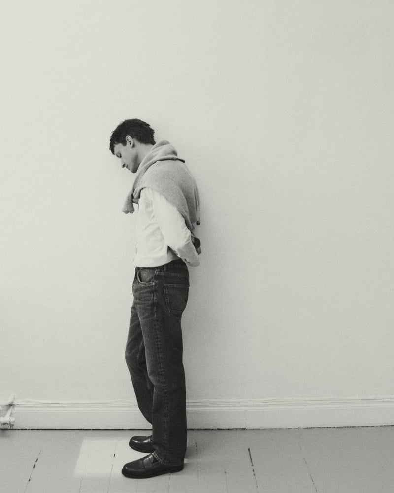 Eddie Klint stands in profile wearing an Oxford shirt and dark jeans with a sweater over his shoulders