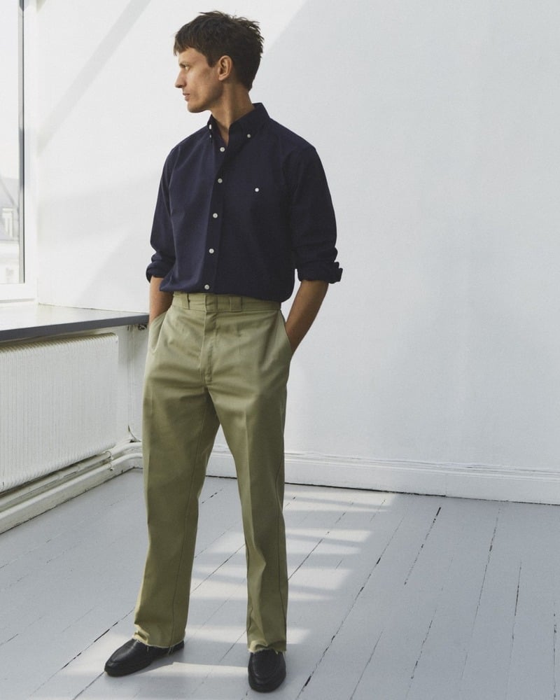 Eddie Klint stands by a window in a navy Oxford shirt and pale trousers