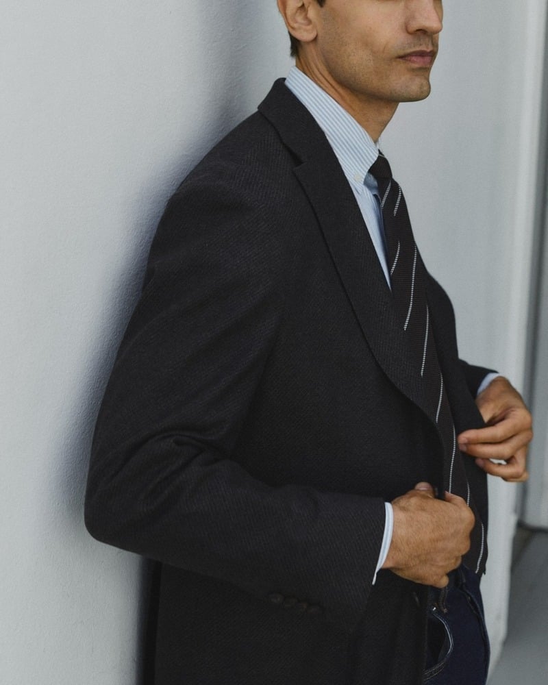 Eddie Klint adjusts a dark tailored jacket over a blue Oxford shirt and striped tie