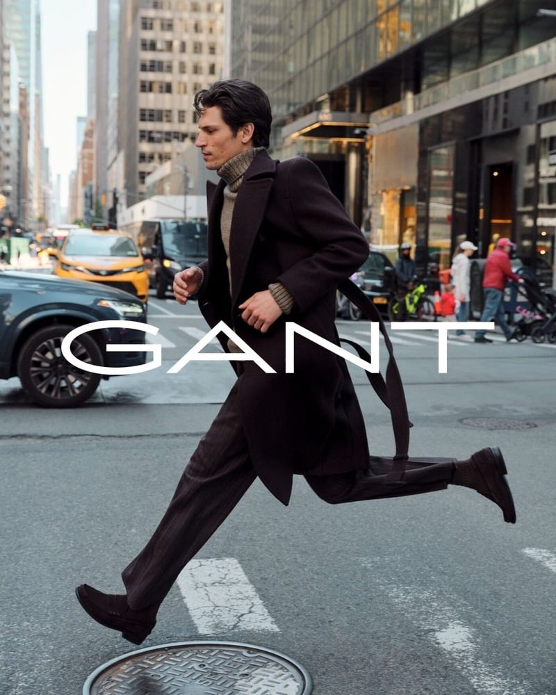 Justin Eric Martin strides across a New York street wearing a dark GANT utility belted coat over a ribbed turtleneck and pinstriped trousers.