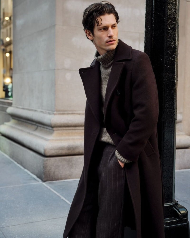 Justin Eric Martin leans against a lamppost on New York's Upper East Side in a dark GANT utility belted coat, ribbed turtleneck, and pinstriped trousers.