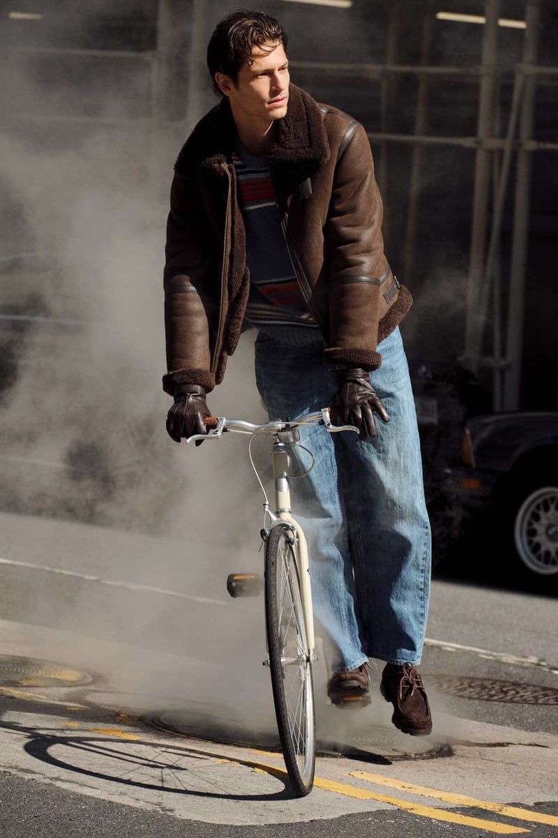 Justin Eric Martin rides a bicycle wearing a brown GANT shearling jacket with a plush collar, striped knitwear, gloves, and relaxed blue jeans.