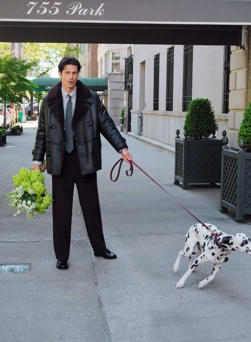 Justin Eric Martin stands with a Dalmatian outside 755 Park Avenue, wearing a dark GANT shearling peacoat over a shirt and patterned tie with dark trousers.