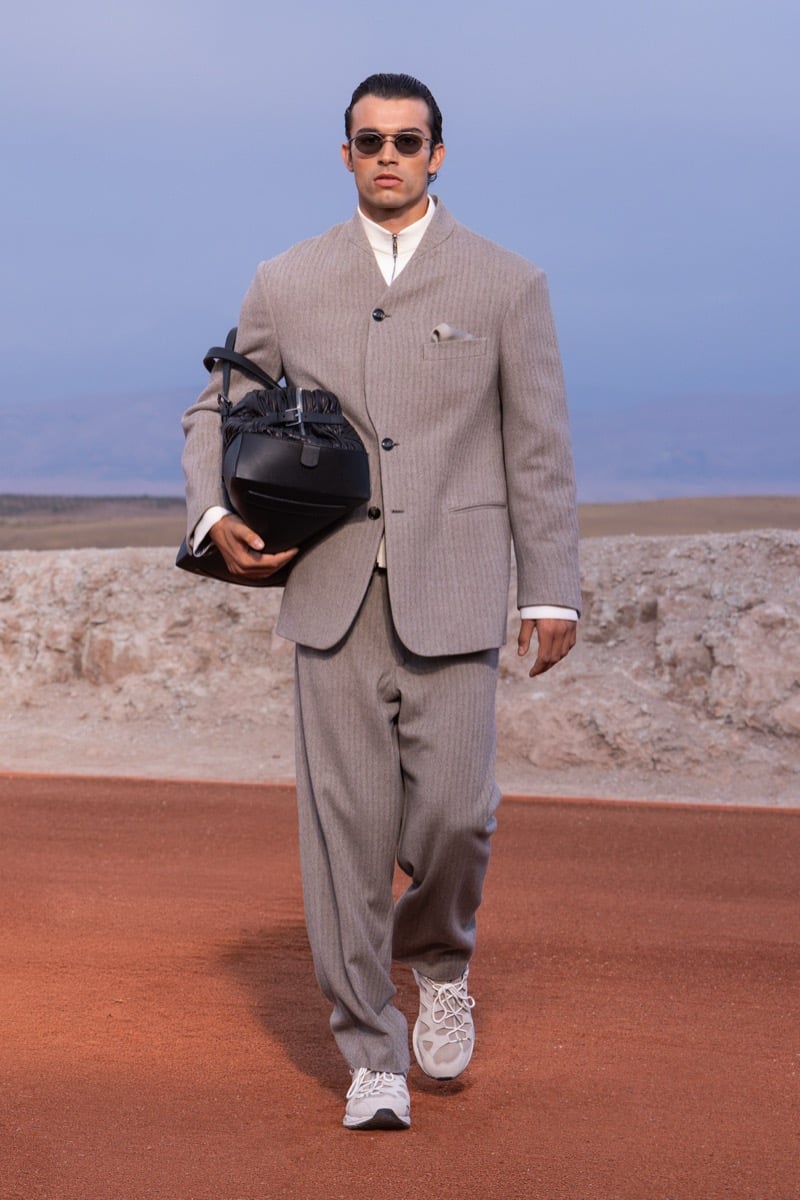 Model wears a taupe tailored suit with a white shirt, sunglasses, and sneakers while carrying a black bag at the Giorgio Armani x Kith fall-winter 2026 show.