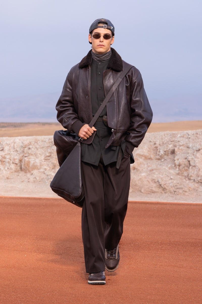 Model wears a dark leather jacket over dark layers and loose trousers with sunglasses and a shoulder bag at the Giorgio Armani x Kith fall-winter 2026 show.
