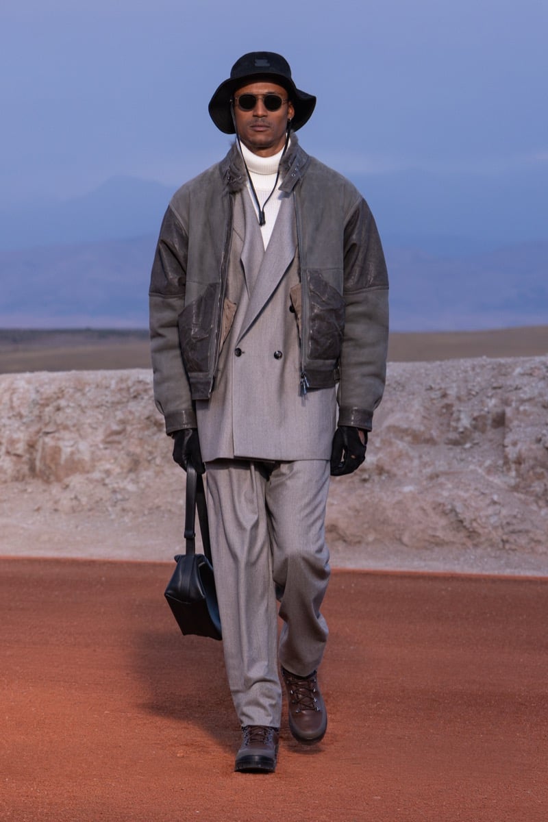 Model wears a charcoal jacket over a gray double-breasted suit with a dark brimmed hat, gloves, and boots at the Giorgio Armani x Kith fall-winter 2026 show.