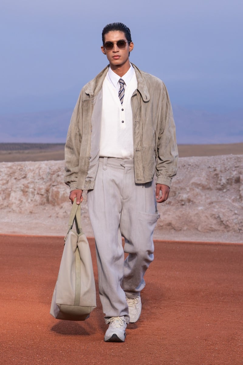 Model wears a light beige jacket, white shirt and tie, loose gray trousers, and sneakers while carrying a large tote at the Giorgio Armani x Kith fall-winter 2026 show.