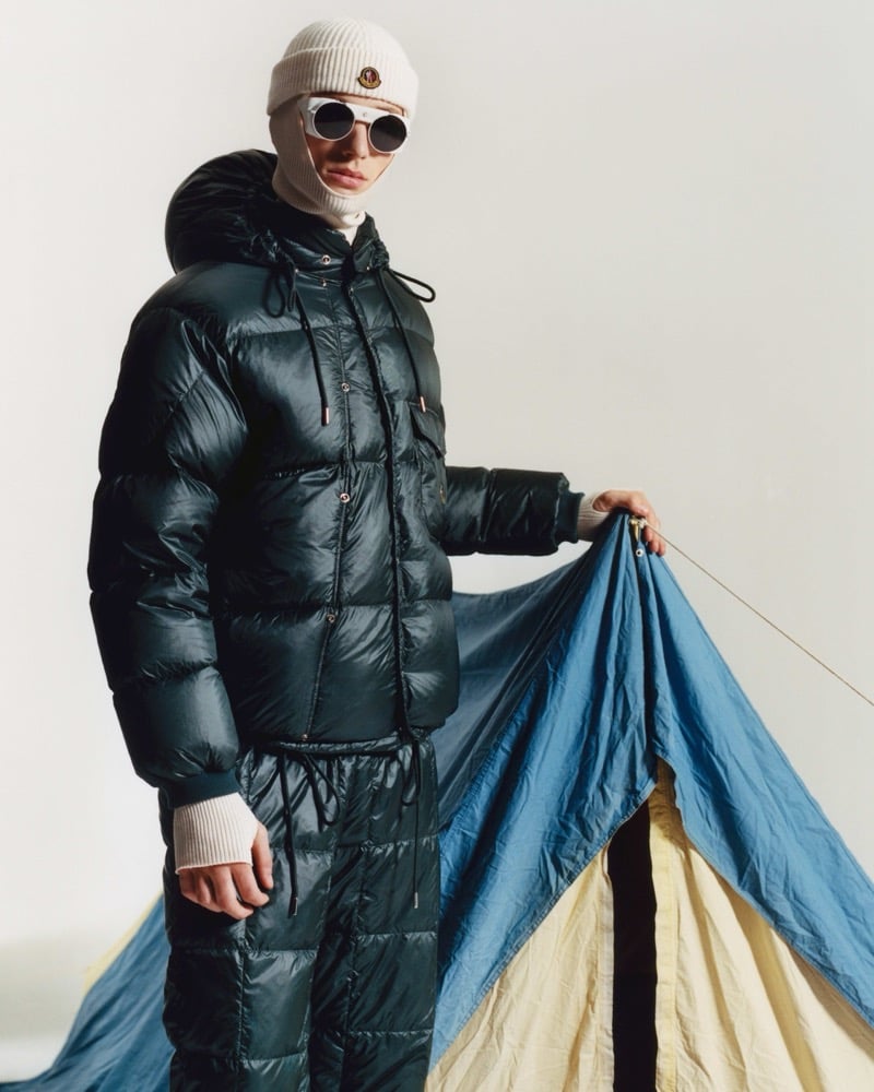 Indiana van't Slot in a knit balaclava, round sunglasses, a glossy dark green puffer jacket and matching quilted trousers, holding up the flap of a blue tent