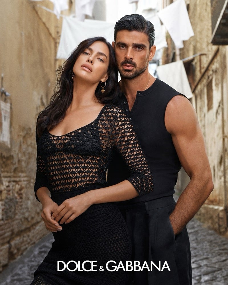 Irina Shayk in an open crochet top and gold heart earrings leaning against Michele Morrone in a ribbed sleeveless henley, in a Naples alley with laundry overhead