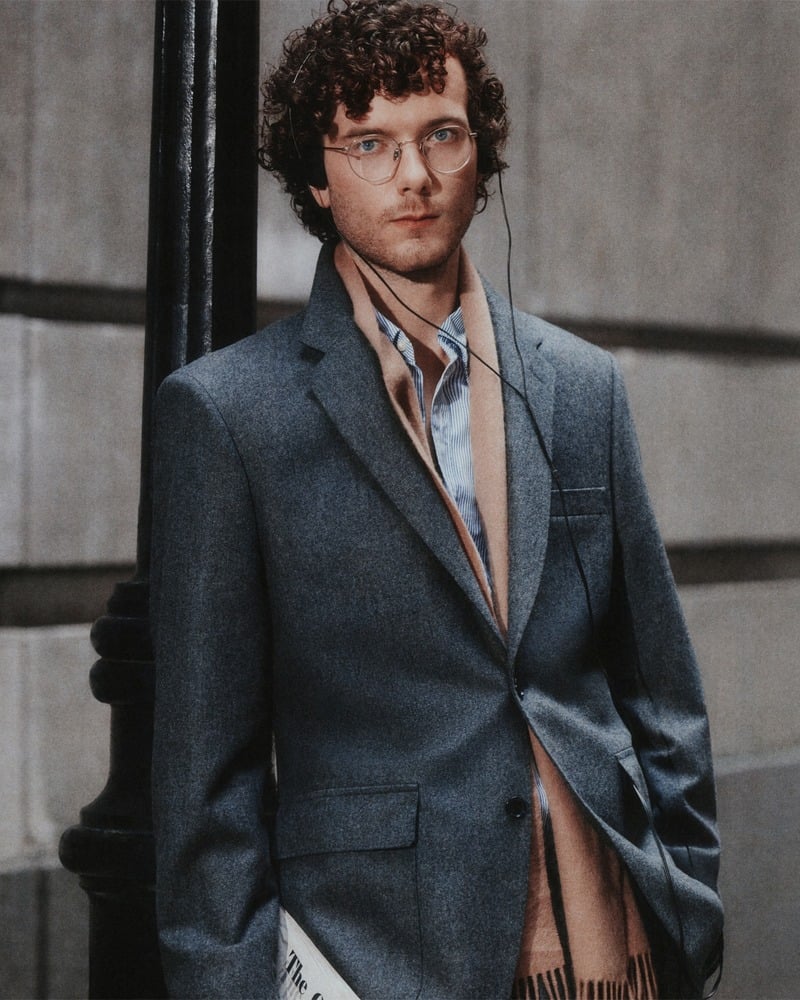 Martin Herlihy wears a gray J.Crew suit with an open striped shirt, scarf, and glasses.