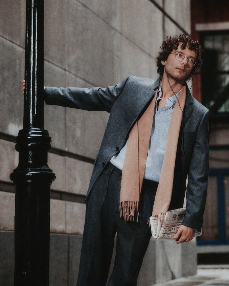 Martin Herlihy leans against a lamppost in a gray suit with a scarf and open shirt.