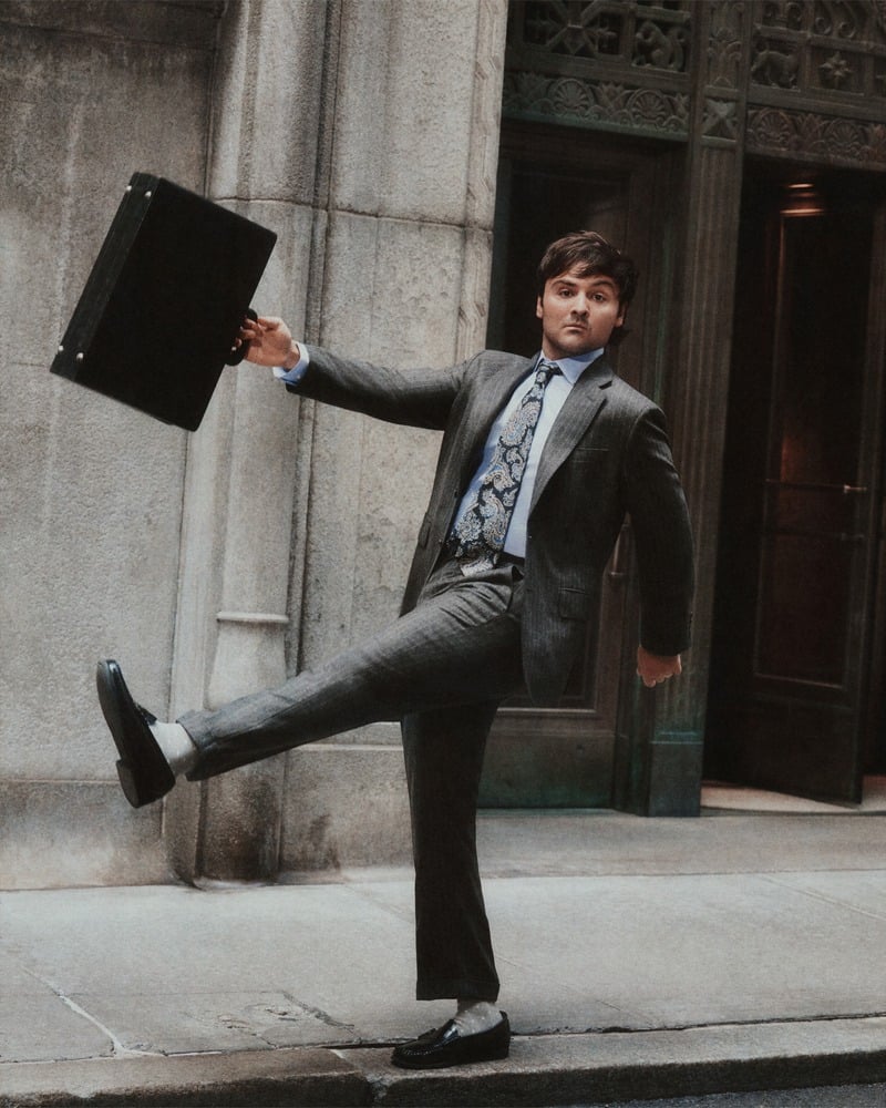 John Higgins kicks one leg into the air while holding a briefcase in a gray suit and patterned tie.