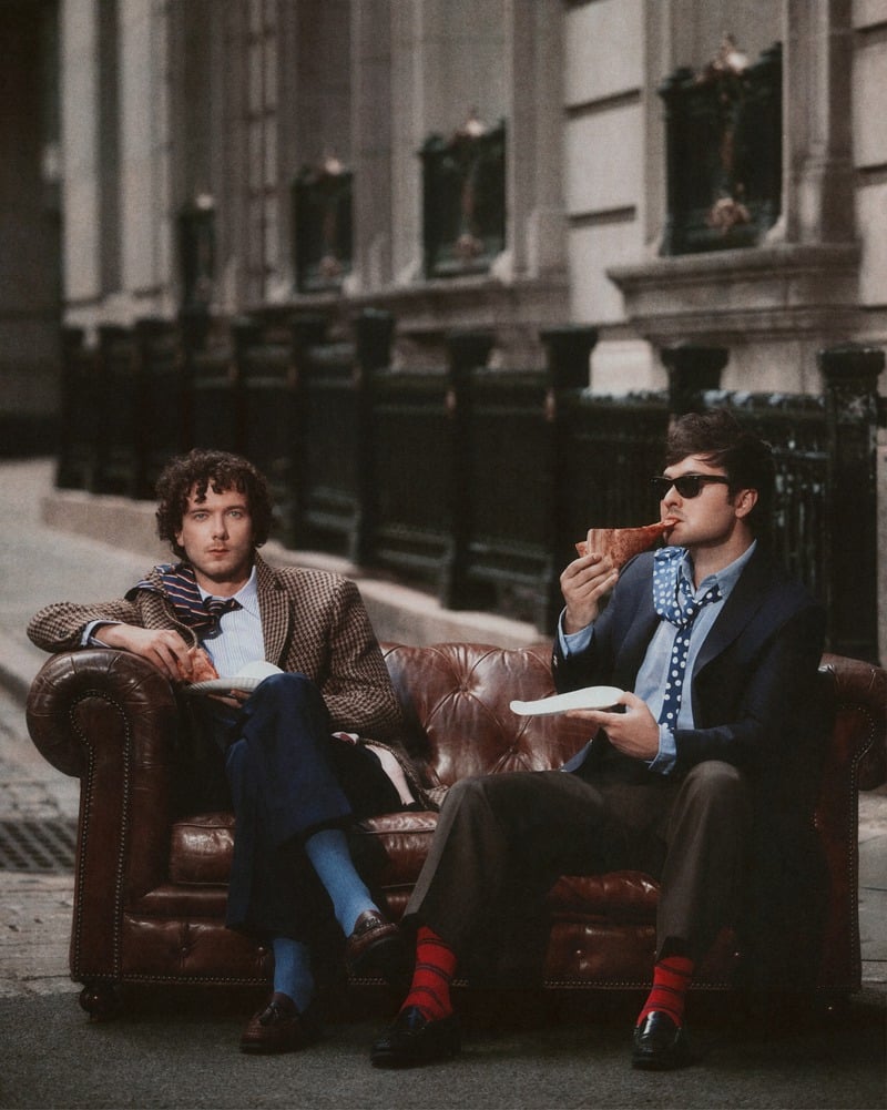 Martin Herlihy and John Higgins sit on a leather sofa on a New York sidewalk with pizza.