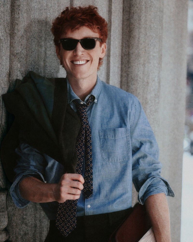Ben Marshall smiles in sunglasses, a blue shirt, and a patterned tie with a dark coat over his shoulder.