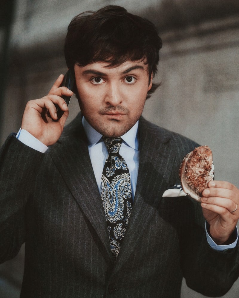 John Higgins holds a pizza slice while speaking on a phone in a gray pinstripe suit and patterned tie.