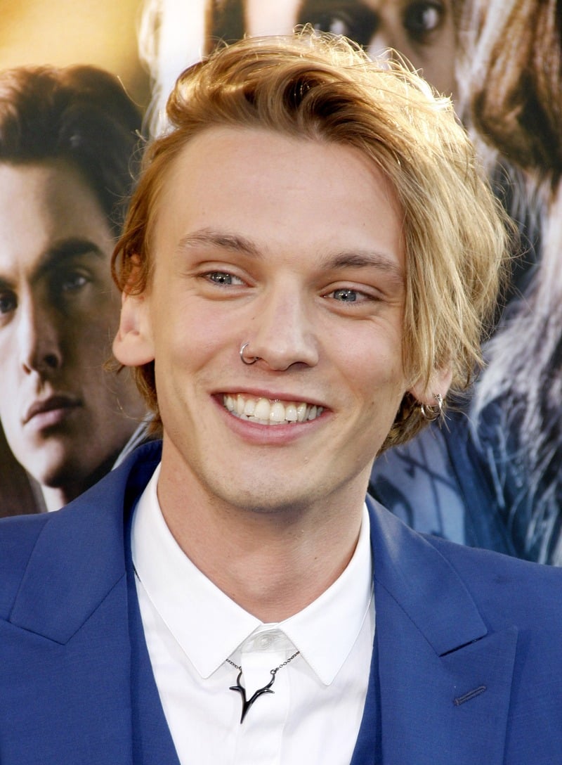 Jamie Campbell Bower smiling with long, side-swept blonde hair, wearing a blue suit and white shirt with a small collar pin.
