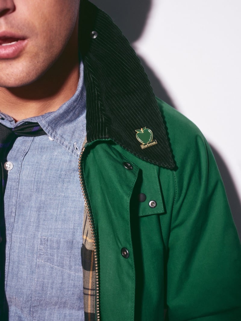 Close-up of the green Beaufort jacket's dark corduroy collar with an enamel apple pin reading Barbour, a brass zip, and tartan lining beside a chambray shirt