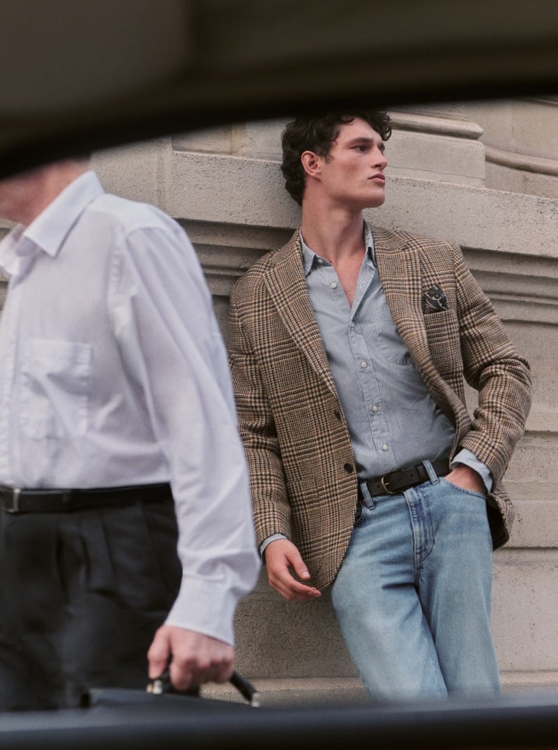 Martin Antech wears a brown glen-plaid sportcoat, striped shirt and faded blue jeans beside a stone wall.