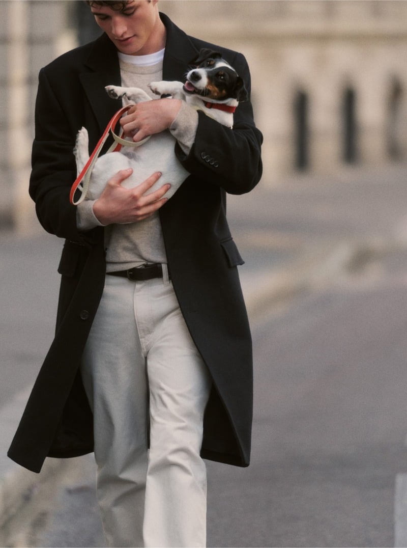 Martin Antech holds a dog while wearing a black topcoat, pale sweater and natural-colored jeans.