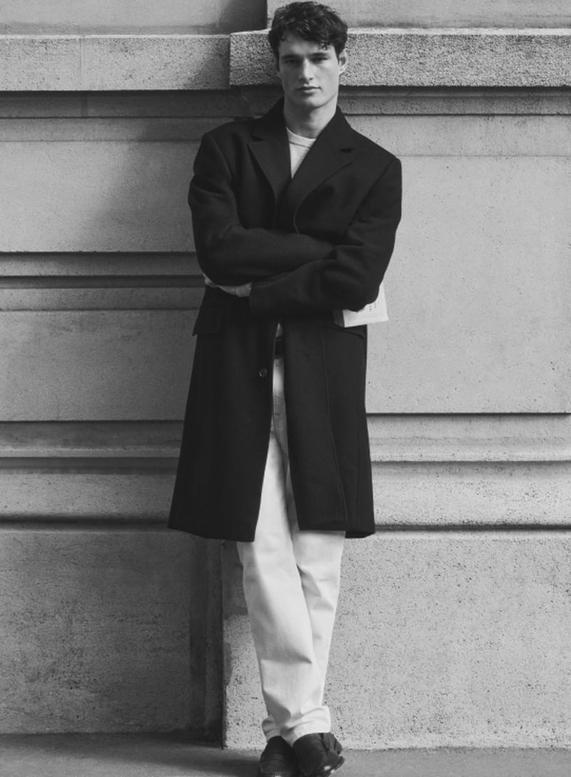 Martin Antech stands against a stone wall in a black topcoat, light trousers and dark loafers.