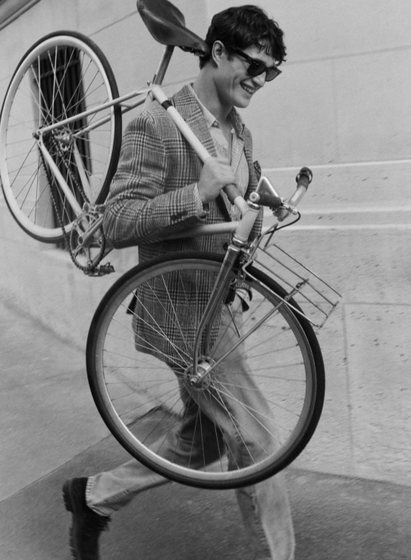Martin Antech holds a bicycle over his shoulder while wearing a glen-plaid sportcoat and sunglasses.