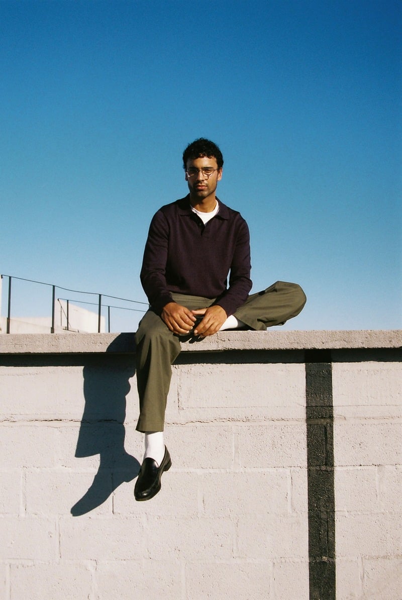 Jy Aoukar sits on a rooftop wall wearing Jimmy Fairly Bellwood glasses, a dark polo, olive trousers, white socks and loafers.
