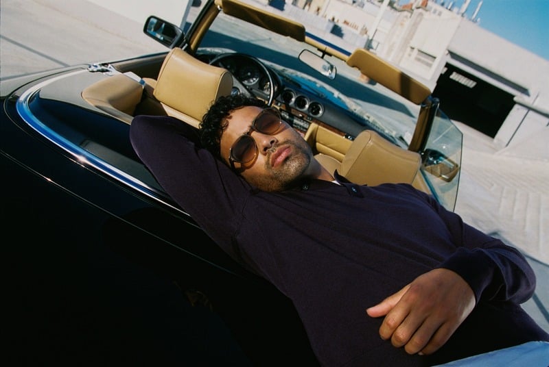 Jy Aoukar reclines against a convertible wearing Jimmy Fairly Harvey sunglasses and a dark polo.
