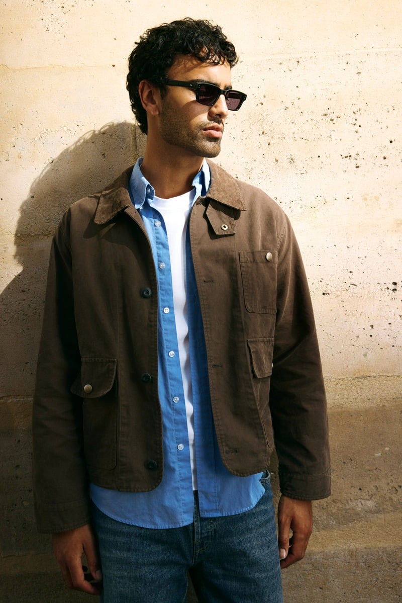 Jy Aoukar wears black Jimmy Fairly Kingsley sunglasses with a brown jacket and blue shirt.