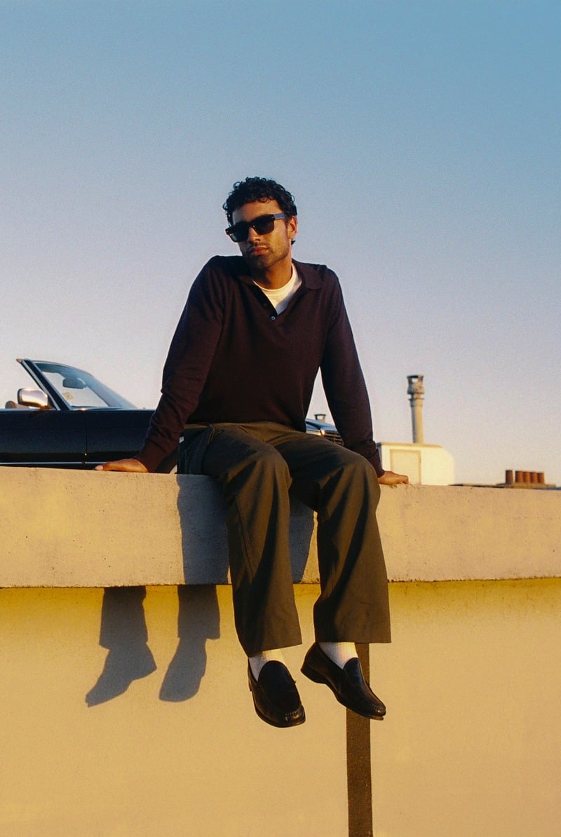 Jy Aoukar sits on a rooftop ledge wearing Jimmy Fairly Lewis sunglasses, a dark polo and olive trousers.
