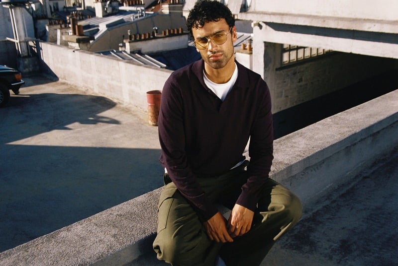 Jy Aoukar sits on a rooftop wearing Jimmy Fairly Parsley tinted metal glasses, a dark polo and olive trousers.