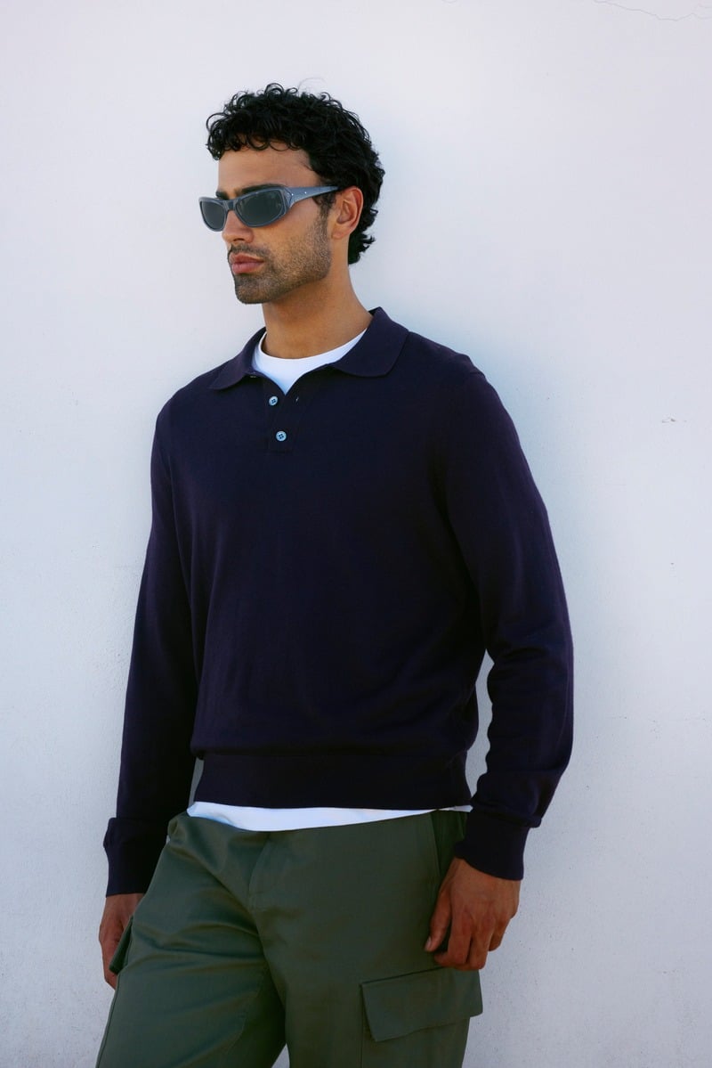 Jy Aoukar wears gray Jimmy Fairly Russell sunglasses with a dark polo and olive trousers.