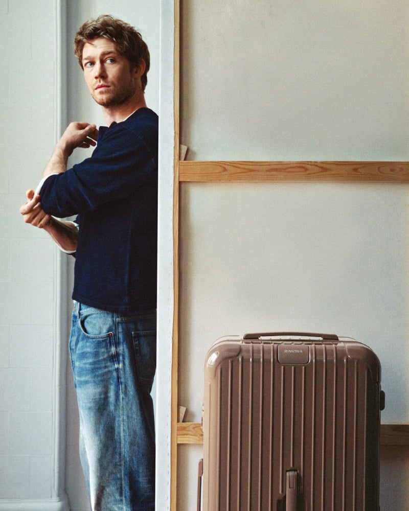 Joe Alwyn leaning against a wooden frame in a knit crewneck sweater and washed denim, adjusting his sleeve, a RIMOWA suitcase standing against the wall beside him