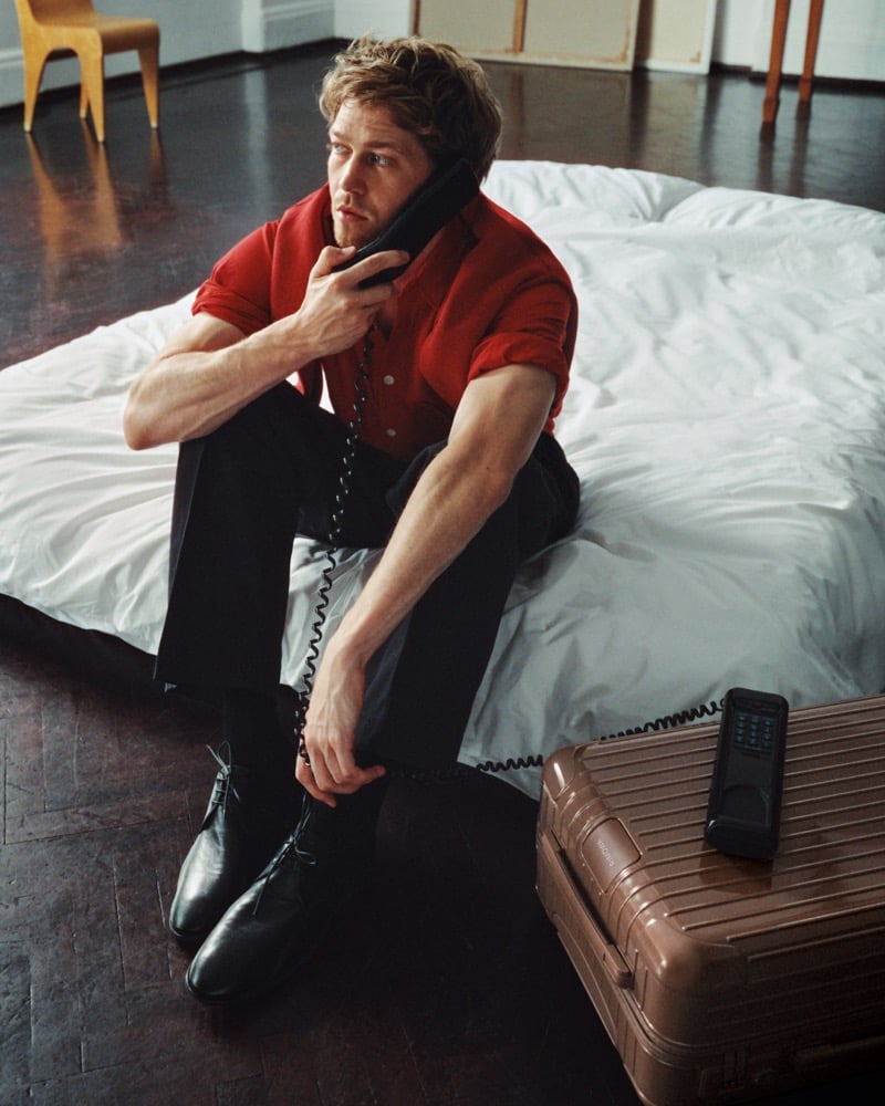 Joe Alwyn sitting on a white mattress on a wood floor in a red short-sleeve shirt, dark trousers, and black lace-up boots, holding a corded phone, a brown RIMOWA suitcase beside him