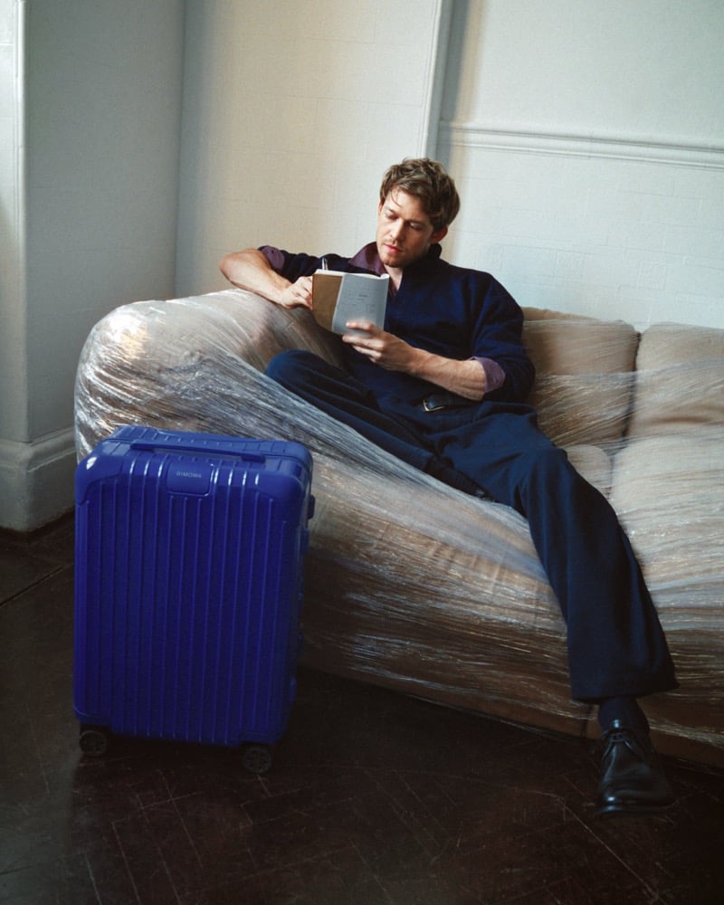 Joe Alwyn reclining on a plastic-wrapped sofa with a notebook in a blue jacket and trousers and black lace-up shoes, an Ultramarine RIMOWA suitcase on the floor at left