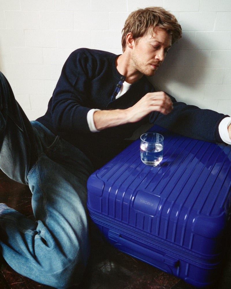 Joe Alwyn seated on the floor against a white brick wall in a knit henley over a white tee and faded jeans, setting a glass of water on an Ultramarine RIMOWA suitcase