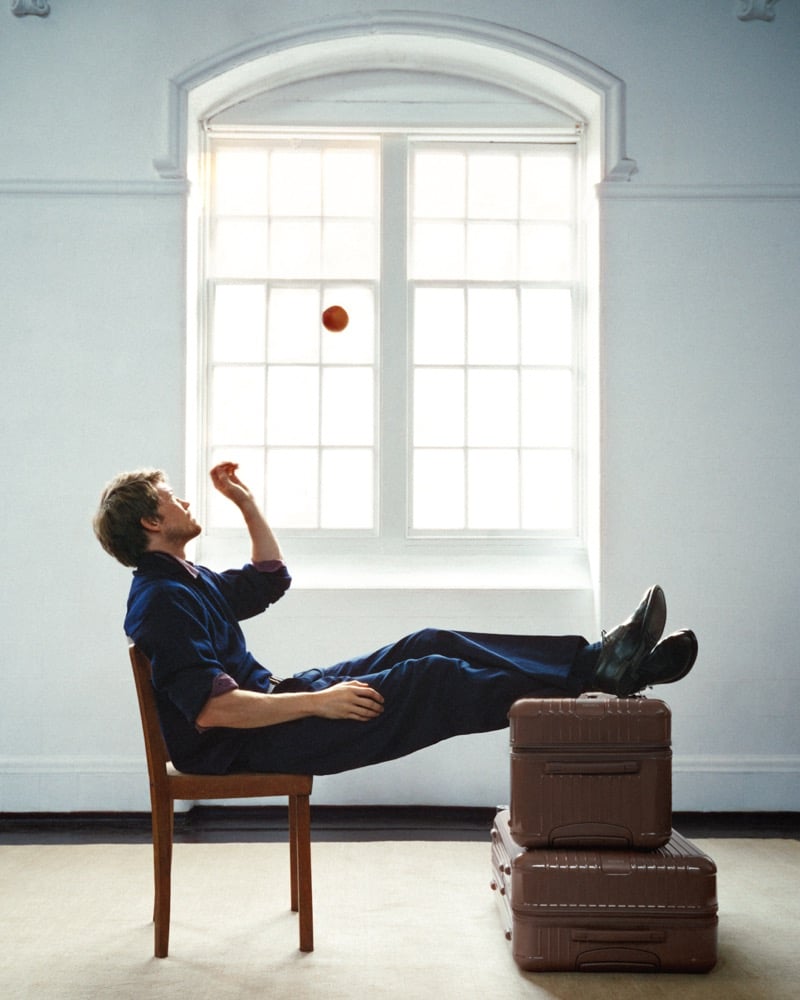 Joe Alwyn seated on a wooden chair with his feet propped on two stacked brown RIMOWA suitcases, tossing an apple in front of a tall arched window