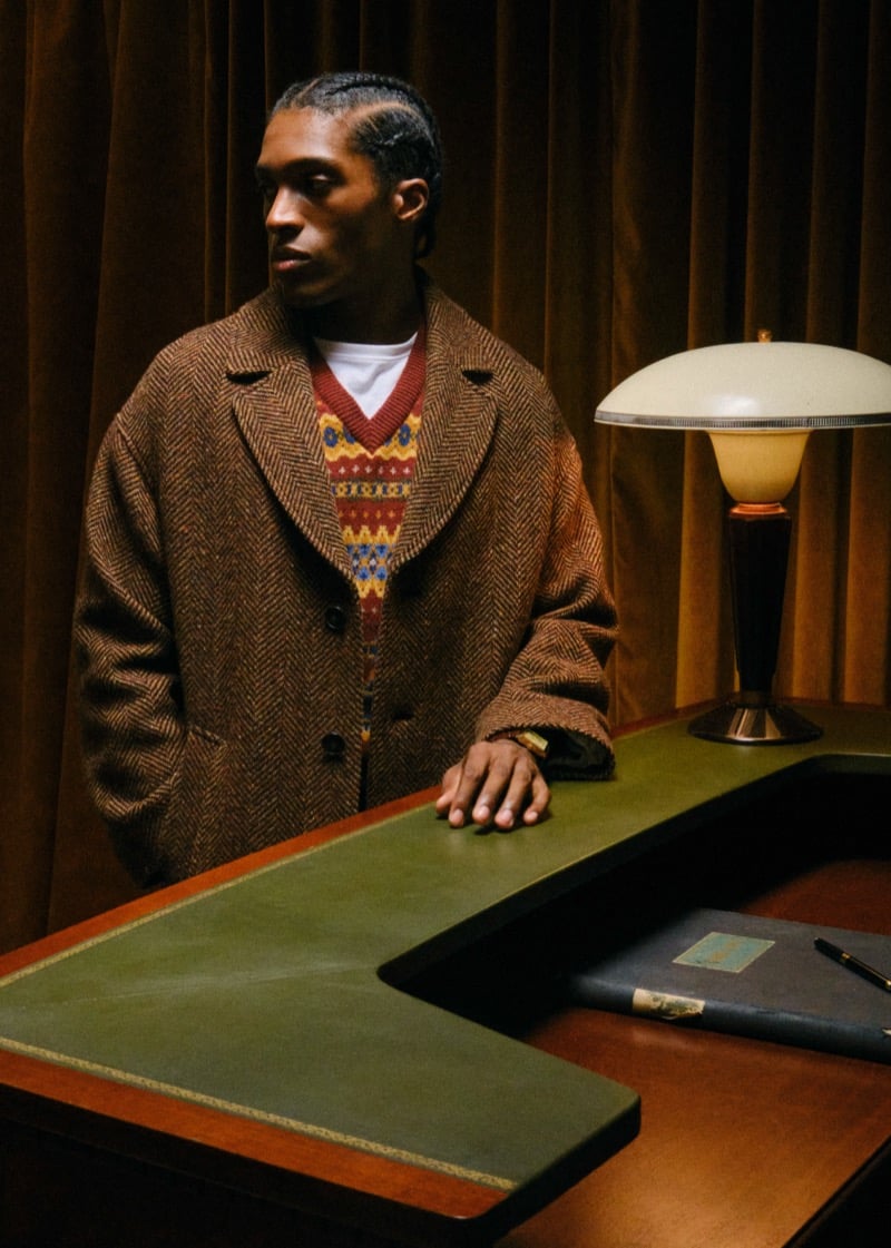 Joe standing at a green leather desk in a herringbone overcoat over a patterned fair isle knit, with a table lamp beside him