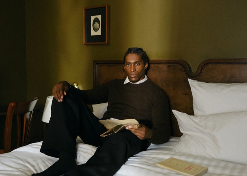 Joe reclining on a hotel bed in a brown sweater with a collared shirt underneath, an open book in his hand