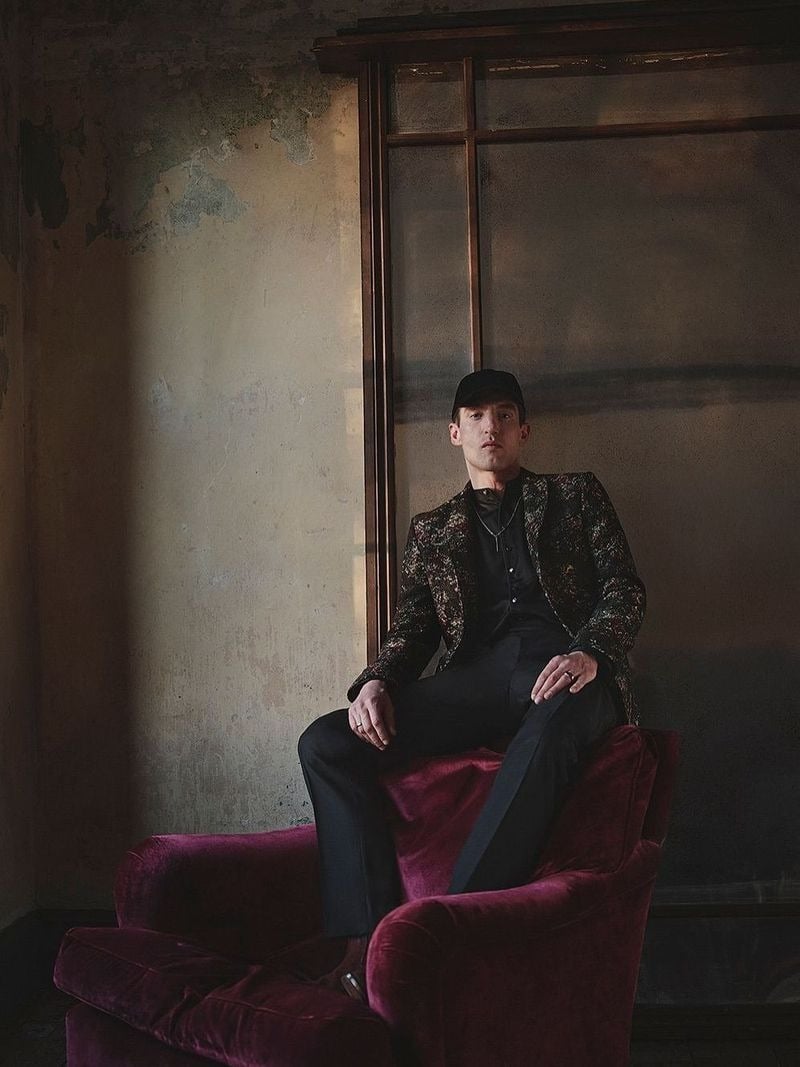 David Trulik sits in a patterned jacket and dark trousers on a red chair for John Varvatos' fall-winter 2026 campaign