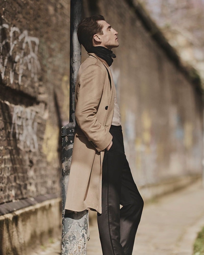 Anatol Modzelewski wears the camel Delmar double-breasted coat beside a stone wall for John Varvatos' fall-winter 2026 campaign
