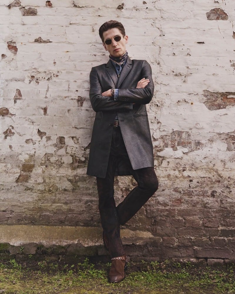 Anatol Modzelewski leans against a brick wall in the Maida coat and sunglasses for John Varvatos' fall-winter 2026 campaign