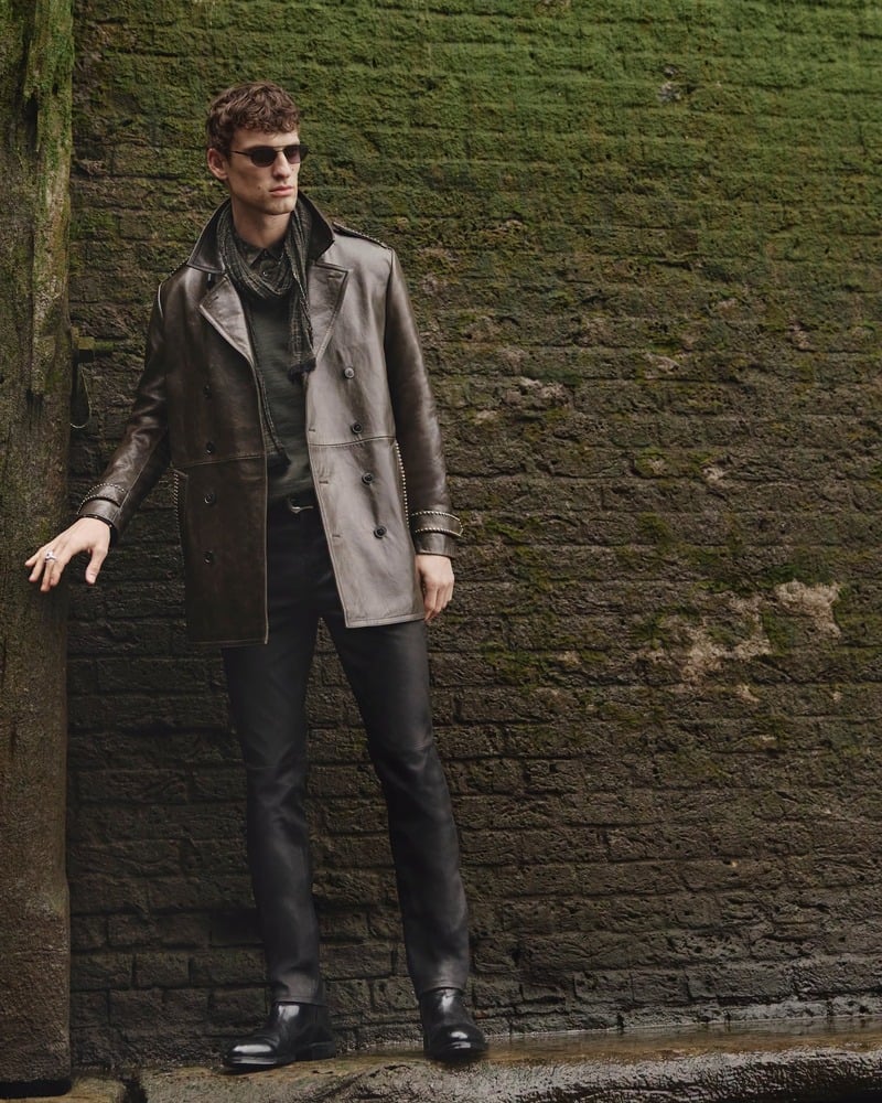 Anatol Modzelewski wears sunglasses and a coat beside a moss-covered wall for John Varvatos' fall-winter 2026 campaign