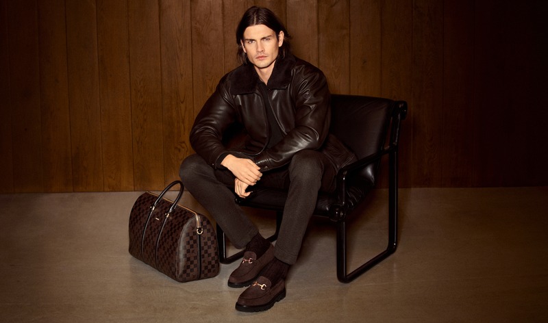 Harrison Page sits in a black chair wearing a dark leather jacket beside a patterned travel bag for JOOP!.