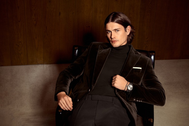 Harrison Page reclines in a black chair wearing a dark velvet jacket and black turtleneck for JOOP!.