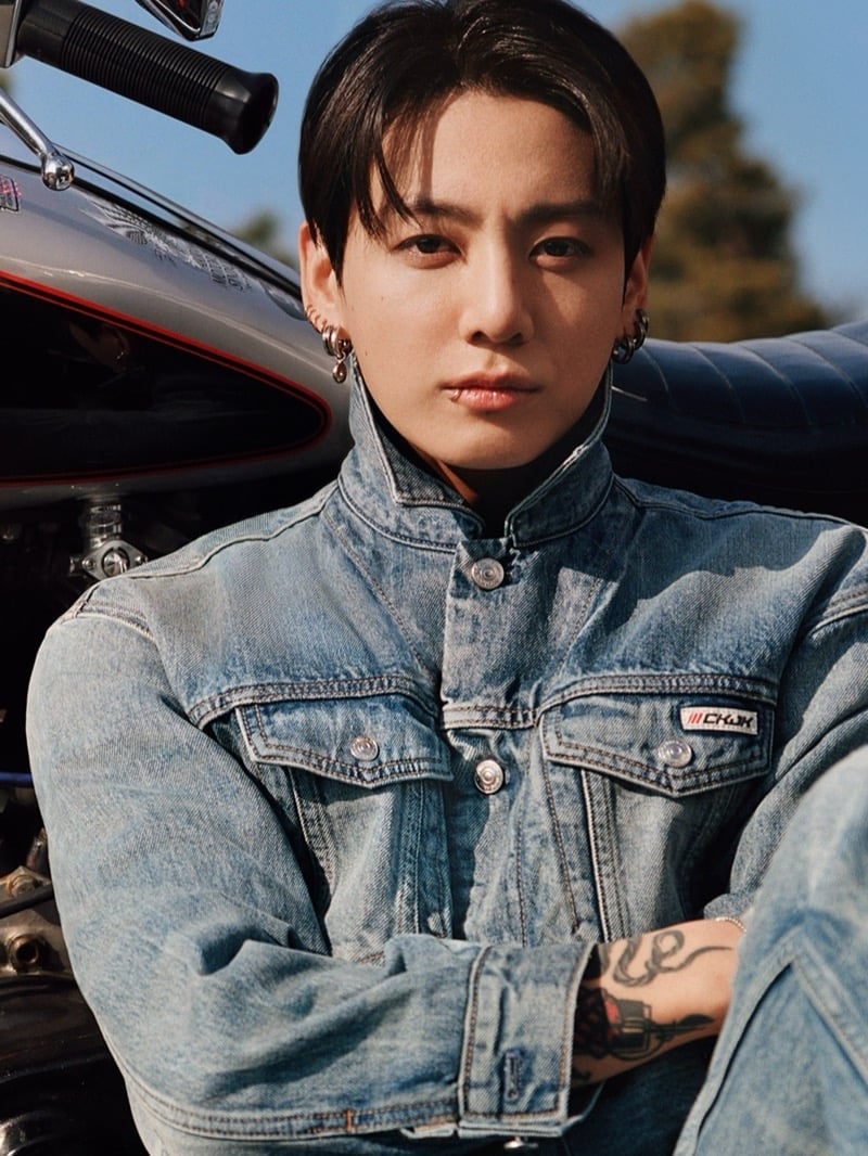 Jung Kook sits beside a black motorcycle in a light-wash denim trucker jacket with a stand collar and red CKJK chest patch.