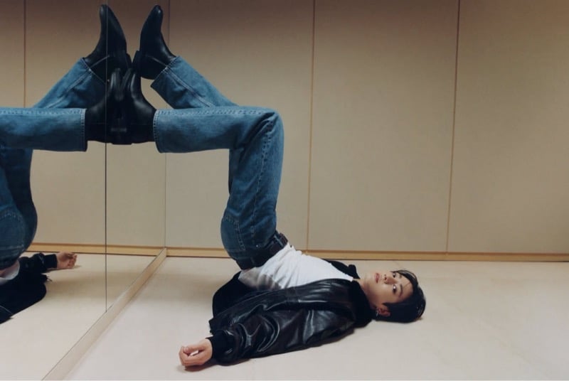 Jung Kook lies beside a mirror with his booted legs raised, wearing jeans, a white tank, and a black leather jacket.