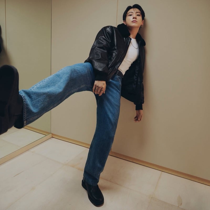 Jung Kook lifts one leg toward the camera in blue jeans, black boots, a white tank, and a leather jacket.